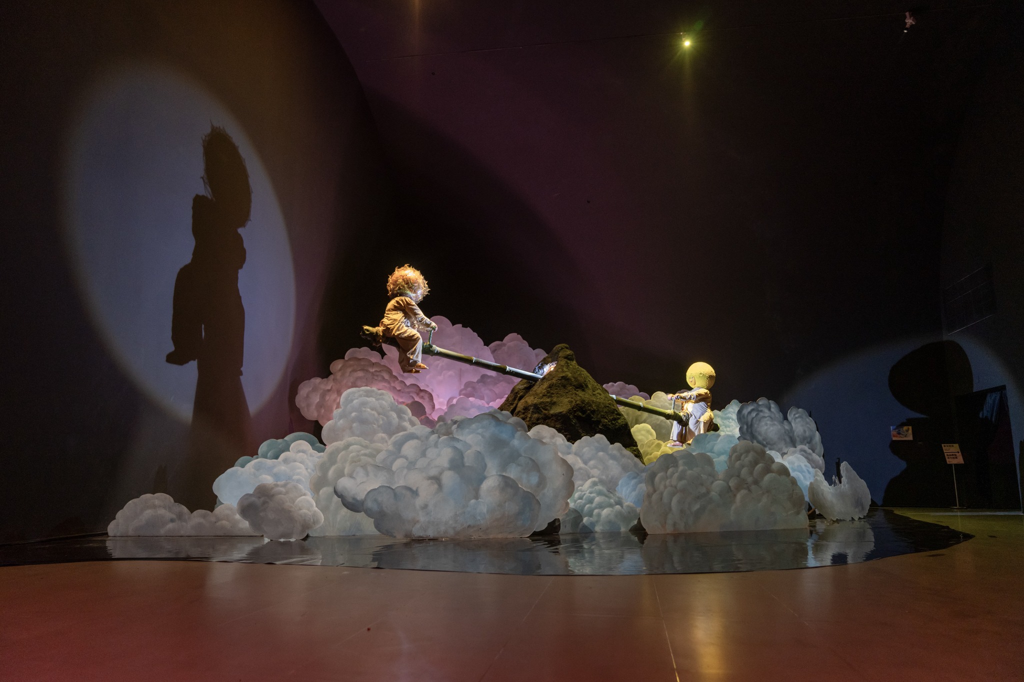 Installation view of the Fuxi: A Feast for the Universe exhibition, showing two figures on a seesaw amid sculpted clouds